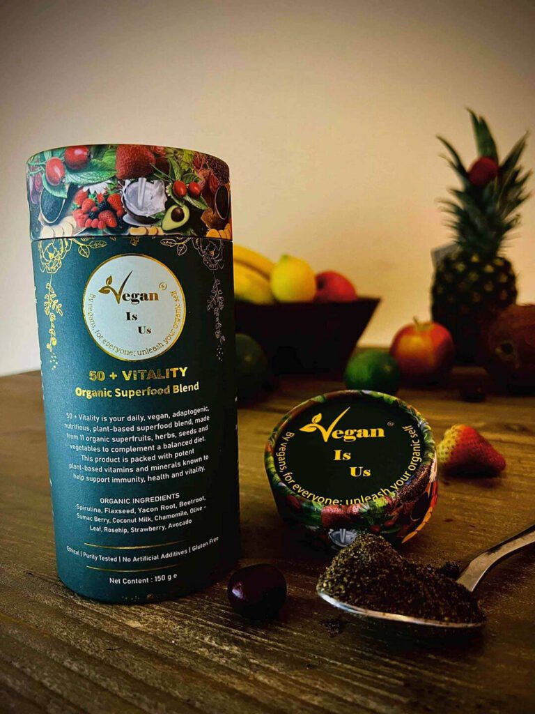 An image of 50 + Vitality organic superfood blend tub that contains Spirulina, Flaxseed, Yacon Root, Beetroot, Sumac Berry, Coconut Milk, Chamomile, Olive Leaf, Rosehip, Strawberry, Avocado as ingredients with tablespoon and fruit bowl on a table,